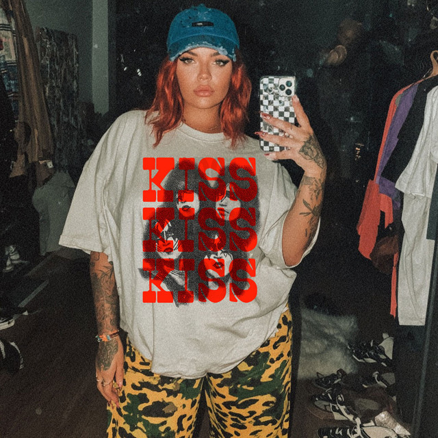 Seventies Band Tee DTF Heat Transfer, a woman with red hair wearing a beige t-shirt with 'KISS' text design, a blue cap, and yellow camouflage pants, taking a mirror selfie in a clothing store.