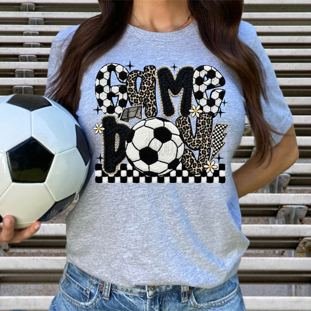Faux Yarn Pattern Soccer Game Day DTF Heat Transfer, image of a woman wearing a t-shirt with soccer ball and sports-themed embroidery design, holding a soccer ball outdoors.