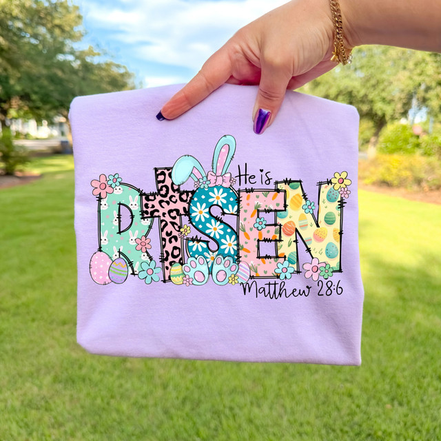 Patterned He Is Risen Easter DTF Heat Transfer, colorful Easter-themed design on a pastel purple clothing item.