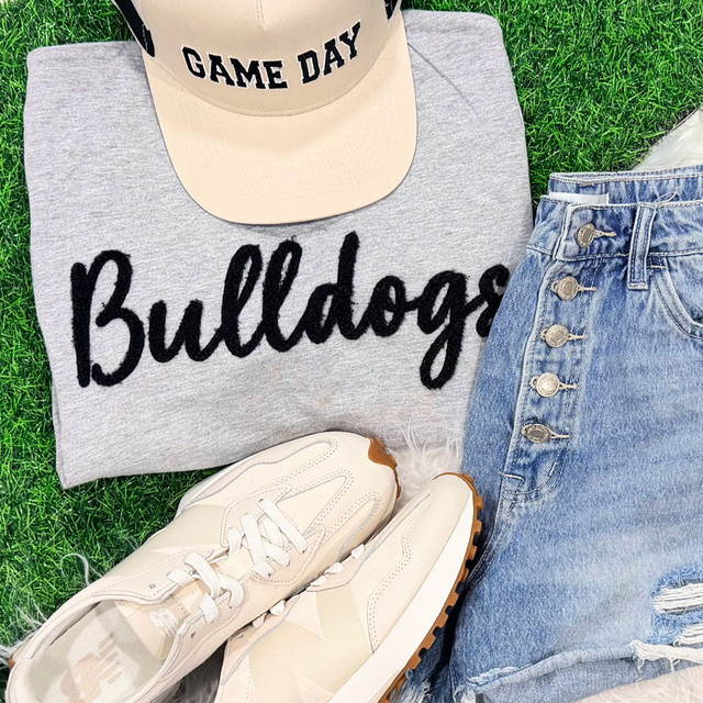 Bulldogs Fluffy Chenille Patch Transfer | Varsity Game Day Patch - Gray sweatshirt with 'Bulldogs' embroidered in black, beige cap with 'GAME DAY' text, distressed denim shorts, and white sneakers on green grass.