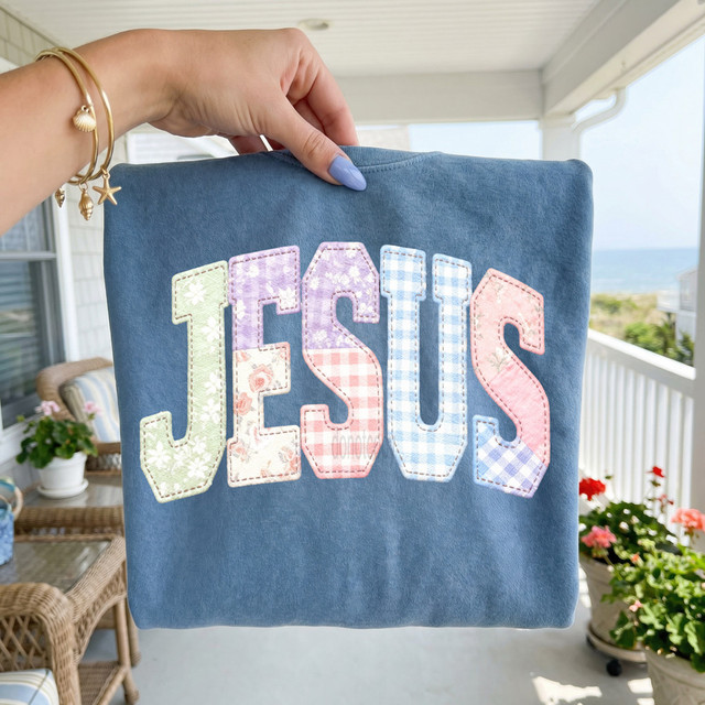 Faux Fabric Patchwork Jesus DTF Heat Transfer, close-up of a hand holding a fabric with colorful patchwork letters spelling 'JESUS' on a blue background, on a porch with outdoor furniture and flowers.