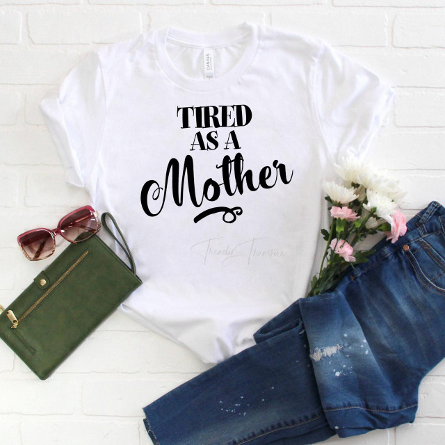Tired as a Mother Sublimation Transfer, white t-shirt with black text design, casual outfit with jeans, sunglasses, a green wallet, and pink and white flowers.