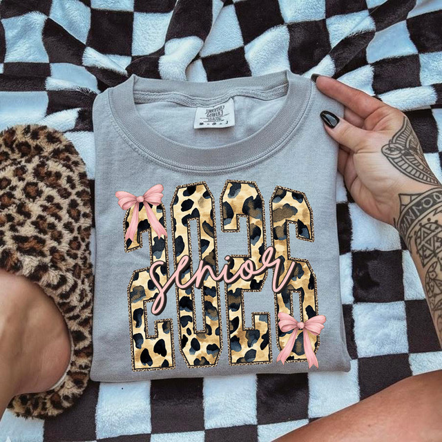 Leopard Senior 2026 Bow DTF Heat Transfer, design of the word 'Senior' with leopard print pattern, pink bows, and cursive 'senior' overlay, on a folded grey t-shirt.