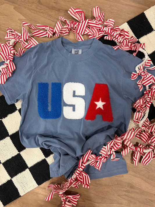 USA Fluffy Patch, decorated T-shirt with 'USA' letters in blue, white, and red with a star, surrounded by red and white striped ribbons on a wooden background.