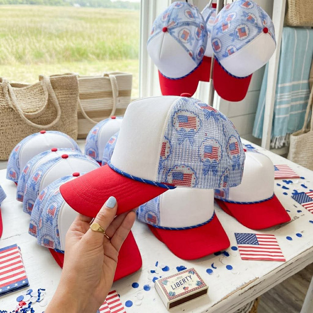 Liberty Cotton Blank Hat, a white and red baseball cap with patriotic American flag patterns, displayed on a table with other similar hats.
