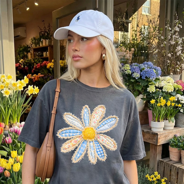 Faux Fabric Patchwork Daisy DTF Heat Transfer, a close-up of a woman wearing a dark gray t-shirt with a large, colorful daisy patchwork floral design on the front, in a flower shop with various flowers in the background.