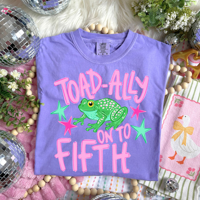 Toad-Ally On To Pink DTF Heat Transfer, colorful graphic T-shirt with green toad and pink text, on lavender fabric surrounded by decorative items.