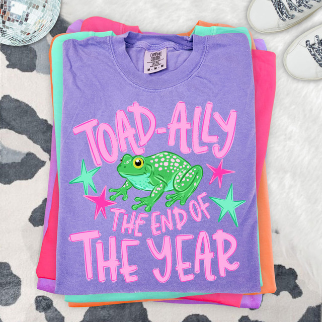 Toad-Ally The End Of The Year DTF Heat Transfer, colorful T-shirt with a frog graphic and pink text on top of stacked colorful clothing.