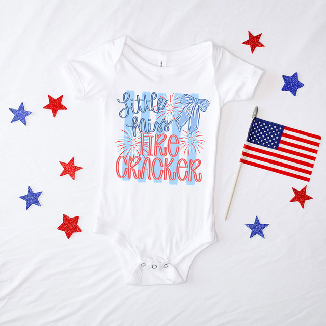 Little Miss Firecracker Bows DTF Heat Transfer, baby onesie with patriotic fireworks design and a blue bow, surrounded by red and blue glitter stars and a small American flag.