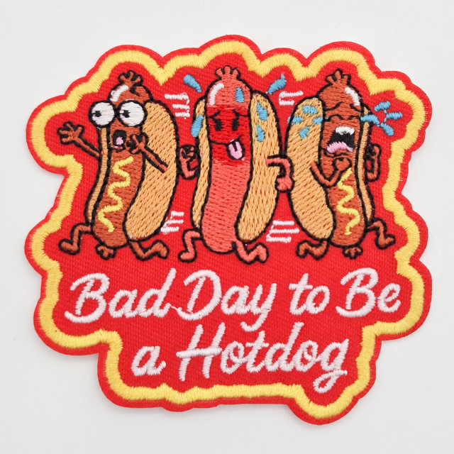 Bad Day to Be a Hotdog, embroidered patch featuring four animated hotdog characters with distressed expressions, against a red background with yellow border, and white text 'Bad Day to Be a Hotdog' underneath.