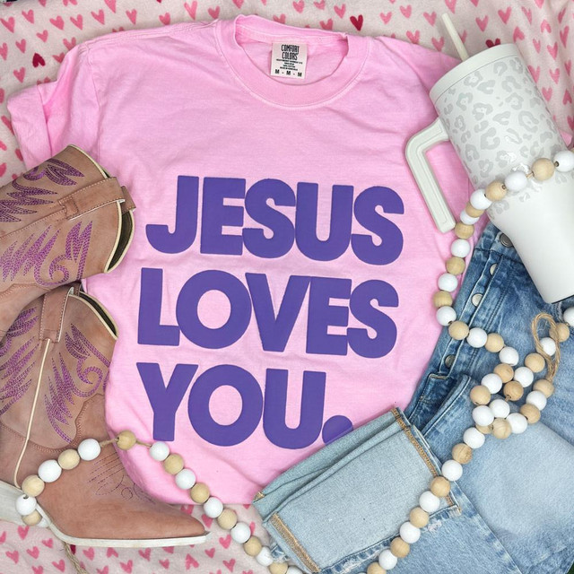 Jesus Loves You Purple PUFF Transfer, product display with pink T-shirt, cowboy boots, mug, beaded necklace, and jeans on a heart-patterned background.