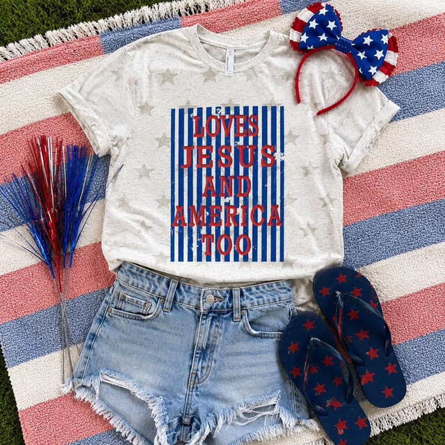 Loves Jesus And America Too Puff Screen Print Transfer t-shirt with patriotic American flag design, paired with denim shorts and red, white, and blue accessories for a themed celebration.