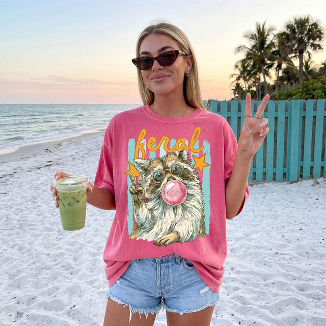 Feral Retro Raccoon DTF Heat Transfer, design of a raccoon wearing sunglasses and blowing a bubble gum bubble, making a peace sign, on a pink T-shirt.