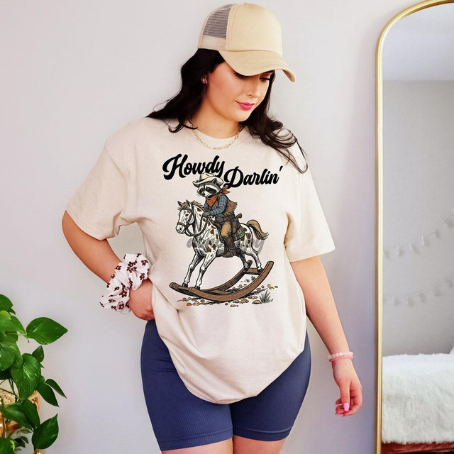 Howdy Darlin' Rocking Horse DTF Heat Transfer, a graphic design of a raccoon riding a rocking horse with bold text, displayed on a woman's cream t-shirt.