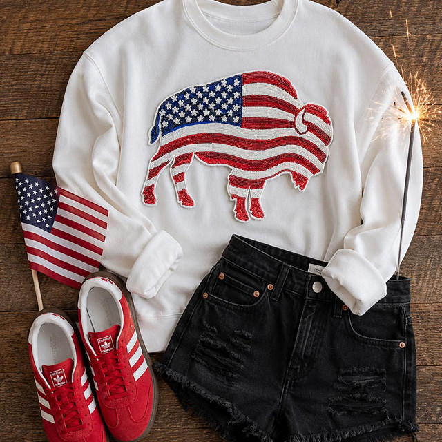 Patriotic Buffalo Sequin Patch, stylish casual outfit with American flag-themed buffalo patch on a white sweatshirt, paired with black distressed shorts, red Adidas sneakers with white stripes, an American flag, and a sparkler on a wooden background.