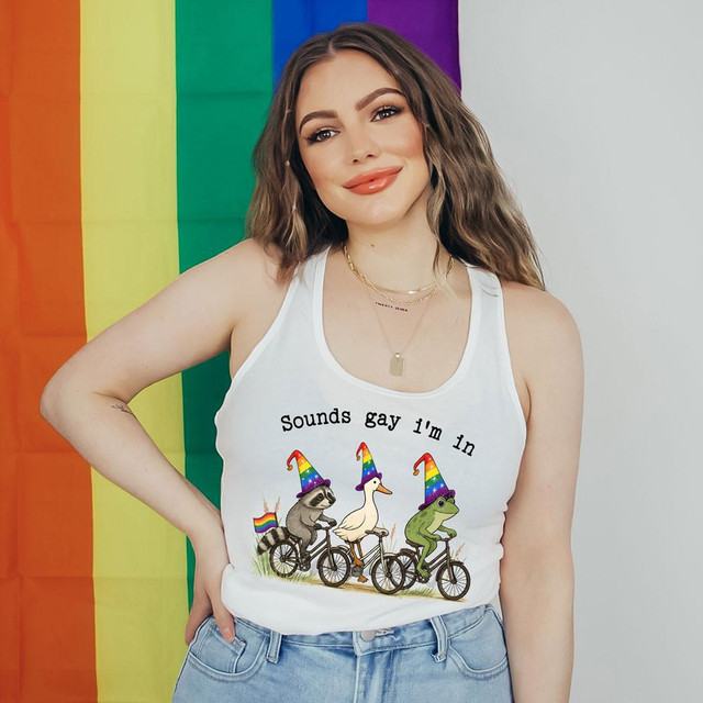 Sounds Gay I'm In DTF Heat Transfer, woman wearing a sleeveless white tank top with colorful cartoon animals riding bikes with rainbow hats and pride flags in the background.