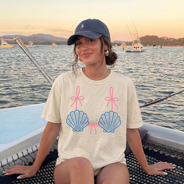 Seashell Bikini Bow Top DTF Heat Transfer, colorful seashell design with pink bows on a cream t-shirt worn by a woman on a boat at sunset.