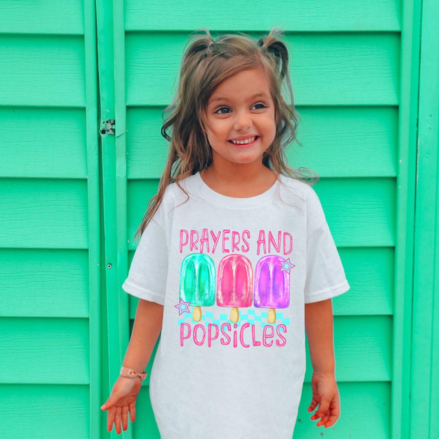 Prayers And Popsicles Pink DTF Heat Transfer, colorful pink, green, and purple popsicle design on a white t-shirt worn by a smiling girl with pigtails standing in front of a mint green fence.