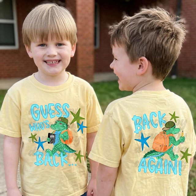 Guess Who's Back... Back Again! BLUE FULL SET DTF Heat Transfer, image of two smiling young boys wearing matching yellow t-shirts with the blue dinosaur design and text on the front and back, outdoor setting with grass and a building in the background.