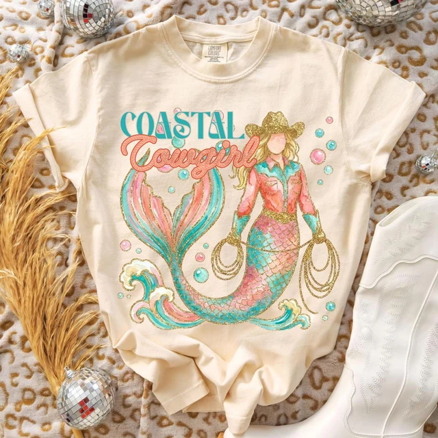 Coastal Cowgirl DTF Heat Transfer, colorful graphic of a mermaid with a cowgirl hat and rope, on a cream-colored T-shirt.