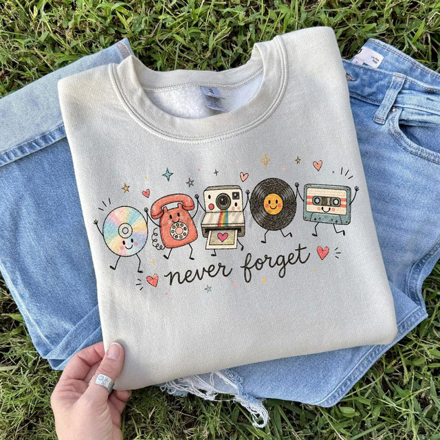 Never Forget Vintage DTF Heat Transfer, colorful cartoon characters of a vinyl record, phone, camera, cassette tape, and the phrase 'never forget' with hearts and stars on a gray sweatshirt.