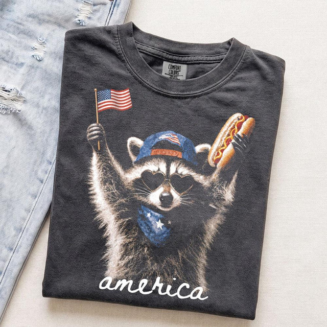 America Raccoon DTF Heat Transfer, a graphic design of a raccoon wearing sunglasses, a patriotic hat, holding a flag and hot dog.