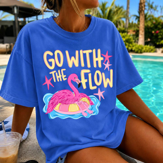 Go With The Flow DTF Heat Transfer, colorful pink flamingo design on a blue T-shirt with text and decorative stars, worn by a woman near a pool with tropical scenery in the background.