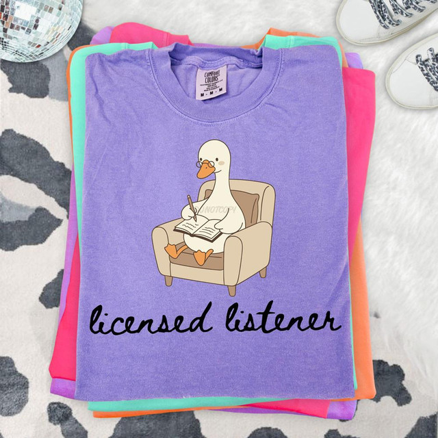 Licensed Listener DTF Heat Transfer T-shirt, purple T-shirt with a cartoon duck sketch sitting in a chair, writing in a notebook, and the text 'licensed listener' printed below.