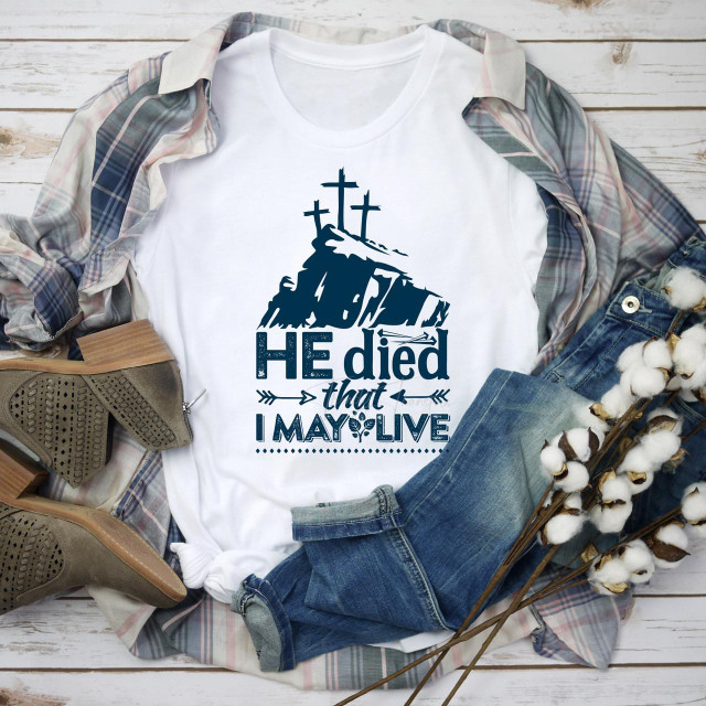 Easter He Died That I May Live Sublimation Transfer, product t-shirt with religious design featuring crosses and a hill, placed on a flat lay with shoes, jeans, plaid shirt, and cotton branches.
