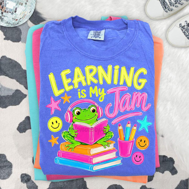 Learning Is My Jam Frog DTF Heat Transfer, colorful graphic t-shirt with a frog reading a book, surrounded by stars and smiley faces, designed for kids or students.