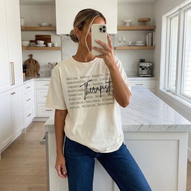 Therapist BLACK DTF Heat Transfer, woman taking a mirror selfie in a bright kitchen wearing a white t-shirt with words printed on it and blue jeans.