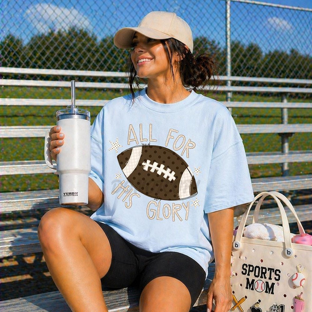 All For His Glory Football DTF Heat Transfer, a woman sitting on bleachers at a sports field wearing a light blue t-shirt with a football graphic and the phrase 'All For His Glory' printed on it.