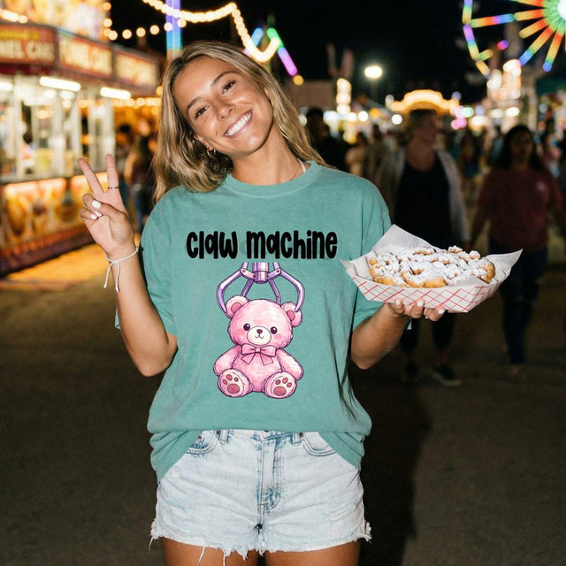 Claw Machine DTF Heat Transfer, a colorful design featuring a pink teddy bear hanging from a claw machine, printed on a teal T-shirt worn by a smiling woman at an amusement park at night.