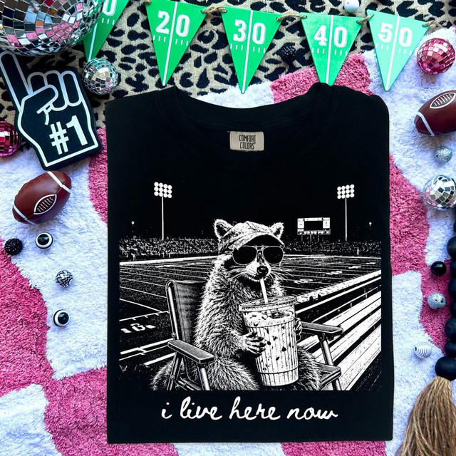I Live Here Now Raccoon DTF Heat Transfer, a black T-shirt featuring a graphic of a raccoon wearing sunglasses sitting on a chair, drinking from a large cup, with a stadium background.
