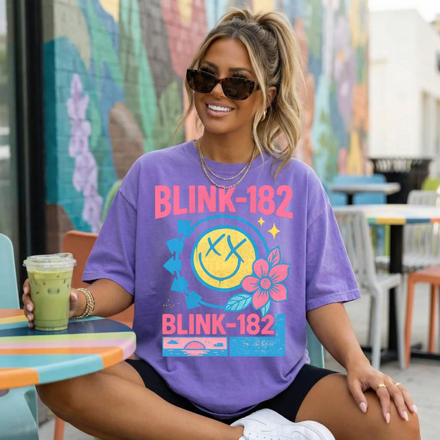 Retro Summer Band Tee DTF Heat Transfer, vibrant purple T-shirt featuring Blink-182 logo and colorful graphic design with smiley face, flowers, and sunset, worn by a smiling woman sitting at an outdoor cafe.