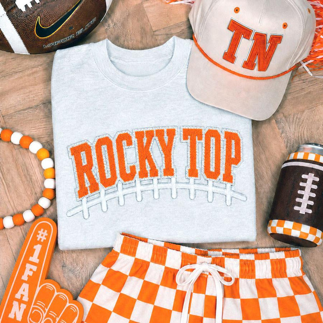 Rocky Top Football Laces Embroidered Chenille Patch, close-up of the patch on a gray sweatshirt featuring orange embroidered text 'ROCKY TOP' with laces design, surrounded by Tennessee Volunteers themed accessories including a hat, football, and themed decorations.