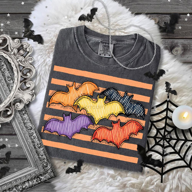 Colorful Bats DTF Heat Transfer, image of a T-shirt with colorful bat designs in orange, yellow, black, purple, and red, set against orange and black striped background with Halloween themed decorations.