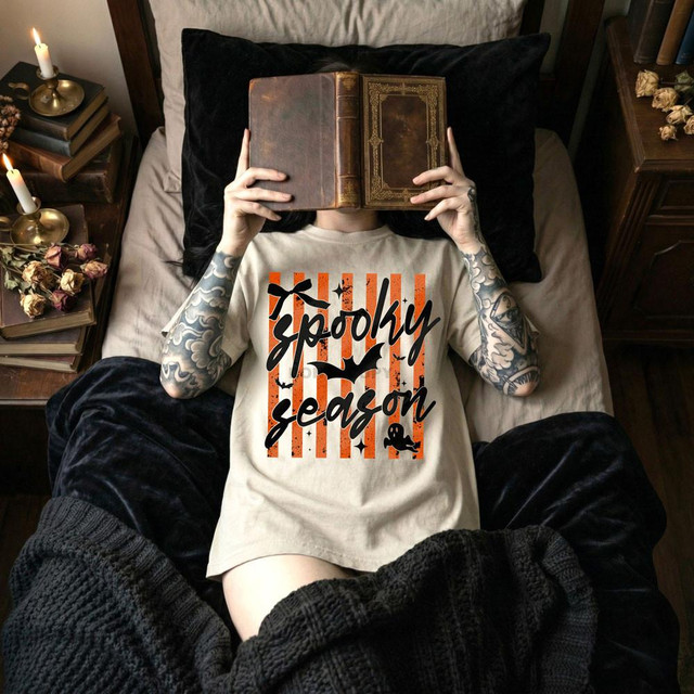 Spooky Season Script DTF Heat Transfer, person lying on bed holding a vintage book, wearing a Halloween-themed T-shirt with orange and black stripes and spooky decorations.