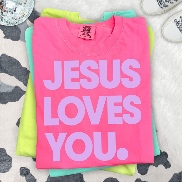 Jesus Loves You LAVENDER DTF Heat Transfer, pink T-shirt with purple text, stacked with green and turquoise shirts on a patterned surface.