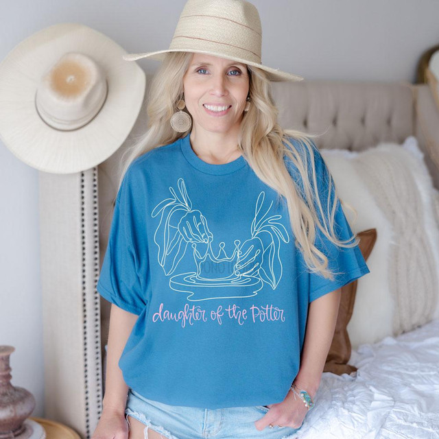 Daughter of the Potter DTF Heat Transfer, woman in blue t-shirt with a line art design of hands holding a pot and text, wearing a wide-brim hat and earrings.