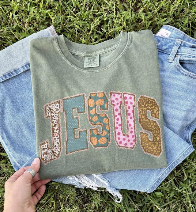 JESUS Faux Applique DTF Transfer | Fall Patchwork Faith Design T-shirt with colorful patterned lettering spelling JESUS on a green background, laid on grass next to light blue denim jeans.