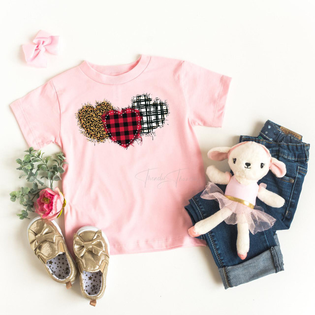 Plaid and Leopard Hearts Youth Screen Print Heat Transfer, pink graphic T-shirt with three heart designs in plaid, leopard, and black and white crosshatch patterns, displayed with plush sheep doll, denim jeans, and girl's shoes with bows and star pattern, for children's fashion and apparel.