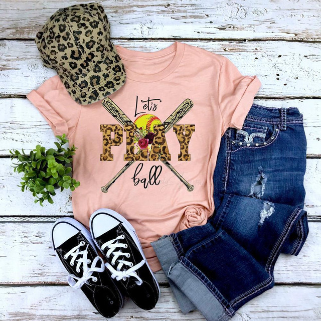 Let's Play Ball Softball Screen Print Heat Transfer T-shirt display with baseballs and bat graphics on pink fabric, paired with jeans, sneakers, a leopard print cap, and a small plant on a rustic wooden background.