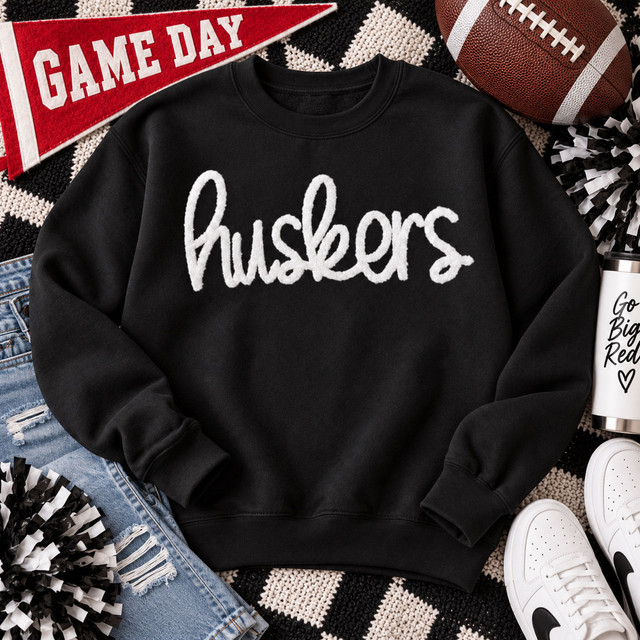 White Huskers fluffy script heat press patch displayed on a black sweatshirt with football game day accessories.