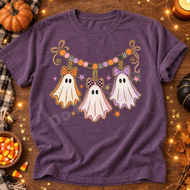 Pastel ghost garland DTF transfer with three bow-topped ghosts in orange, pink, and purple strung beneath a beaded candy-colored banner with stars and glitter accents Shown on a Violet t-shirt.