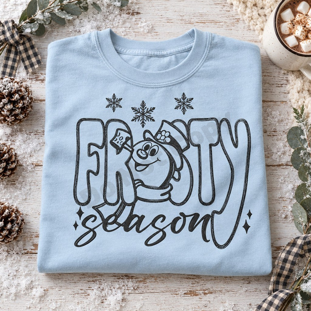 Frosty Season DTF transfer with cartoon snowman in top hat peeking through chunky bubble letters, snowflakes, and brush-script lettering Shown on a Ice Blue t-shirt.