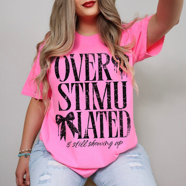 Overstimulated and still showing up DTF transfer with distressed black block lettering and a hand-drawn bow on a pink shirt