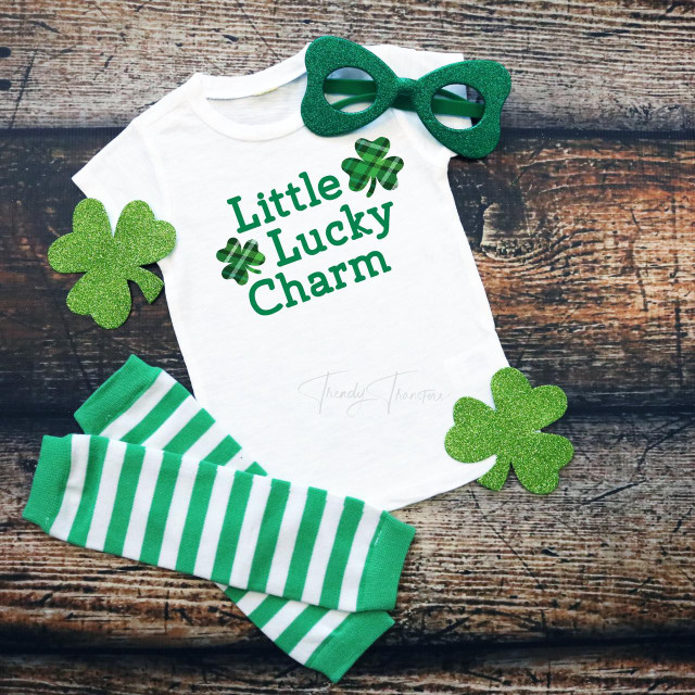 Little Lucky Charm PLAID INFANT Screen Print Transfer, a white infant T-shirt with green text and shamrock accents, surrounded by green glitter shamrock cutouts, green and white striped socks, and green glitter glasses on a rustic wooden background.