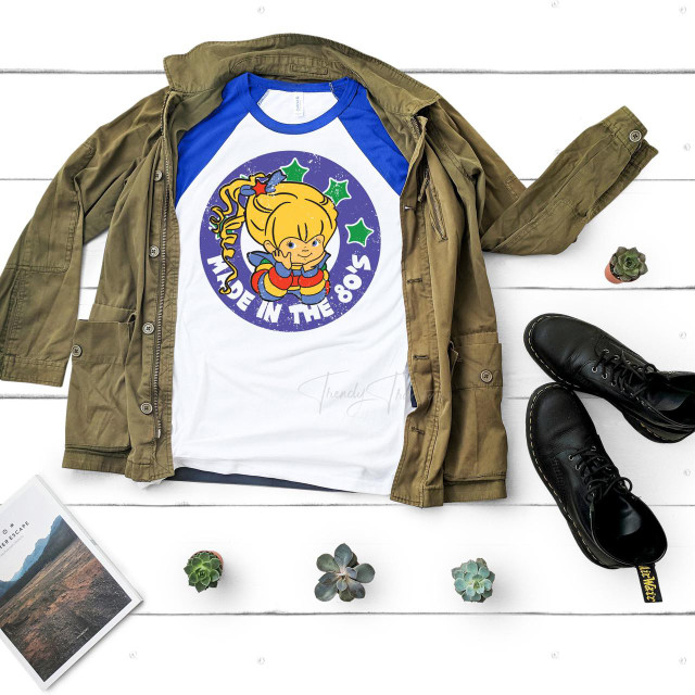 Made in the 80s Rainbow Brite Sublimation Transfer, graphic design featuring a colorful cartoon girl with stars, on a T-shirt with a navy blue and white sleeves, laid on a white wooden surface with a khaki jacket, black boots, small potted plants, and a magazine.
