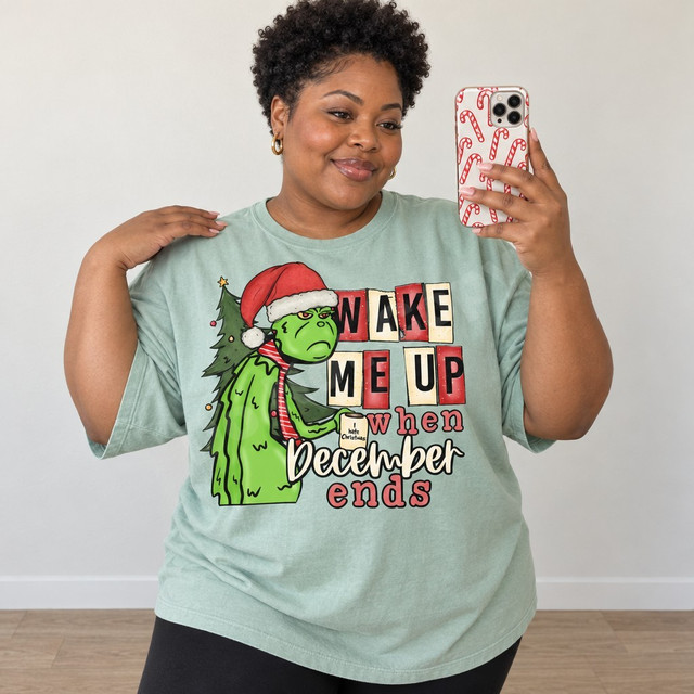 Grumpy green Grinch-inspired character in a Santa hat and candy-striped tie holding an 'I Hate Christmas' mug beside a decorated tree, with bold text reading 'Wake Me Up When December Ends' DTF transfer design Shown on a Seafoam t-shirt.
